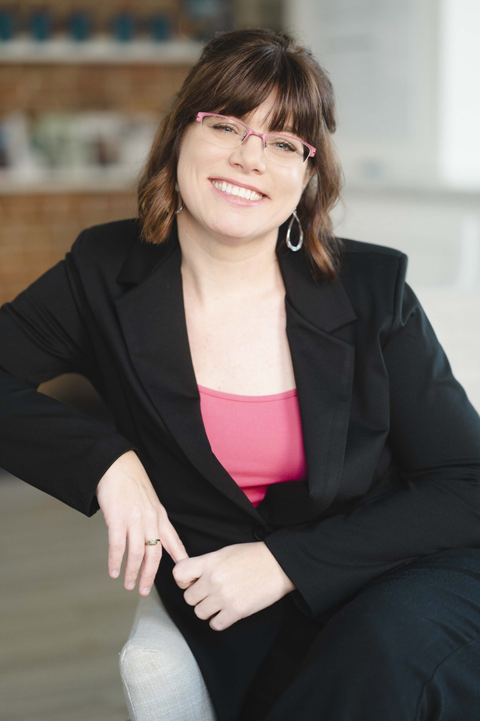 Portrait de Marie-Josée R. Roy, une femme souriante à lunettes roses, portant un veston noir sur un haut rose.
