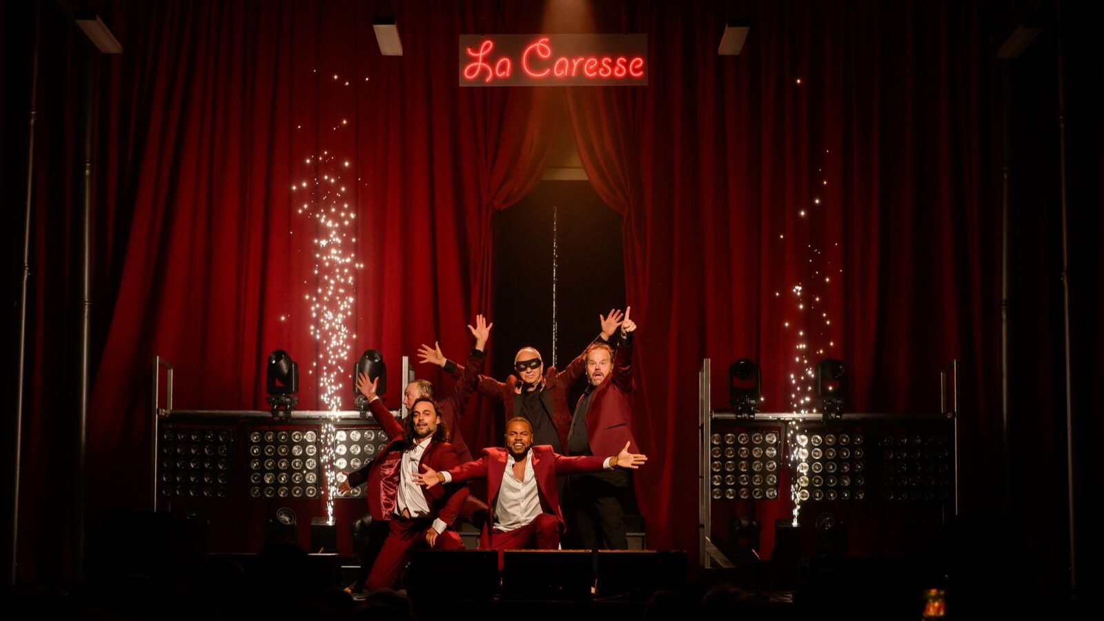 Les acteurs de la pièce Ladies Night vêtus de vestons et pantalons rouges sur chemises blanches, accroupis en petit groupe, devant un rideau rouge sur scène, avec un néon écrit La Caresse haut derrière eux.