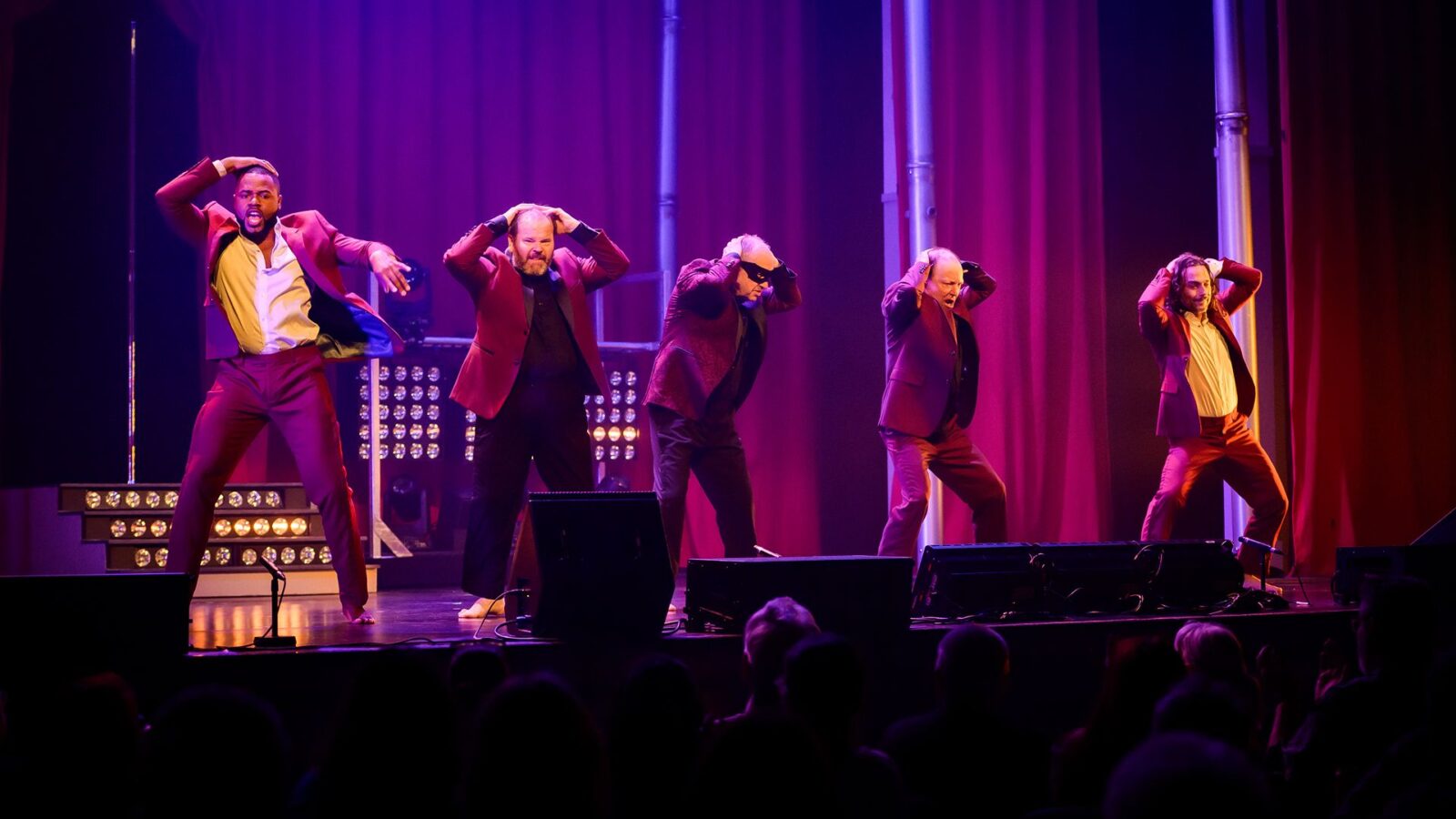 Les acteurs de la pièce Ladies Night, l'un à côté de l'autre, dans des habits rouges, en train de danser de façon sexy, devant un rideau rouge et dans un éclairage bleu-rouge sur scène