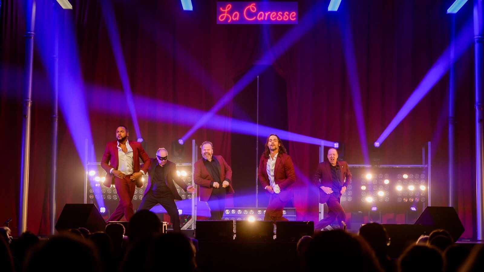 Les acteurs de la pièce Ladies Night, l'un derrière l'autre, dans des habits rouges, en train de danser de façon sexy, devant un rideau rouge et dans un éclairage bleu-rouge sur scène, avec autour d'eux des faisceaux d'éclairage bleus et des ampoules illluminées blanches