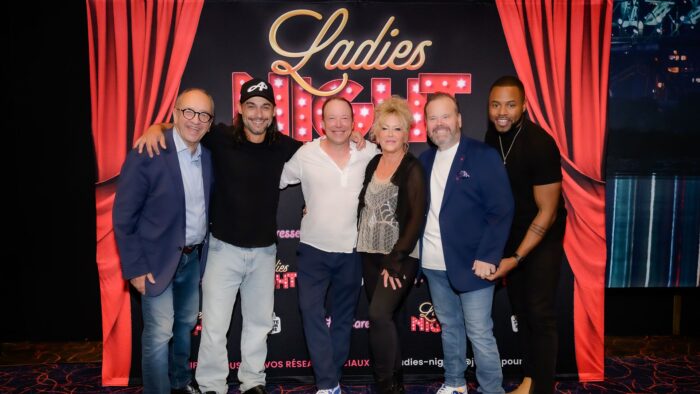 Les comédiens de la troupe Ladies Night, Marcel Leboeuf, Pierre-Paul Alain, François Chénier, Frédérike Bédard, Michel Charette et Lyndz Dantiste devant le logo beige et rouge de la pièce Ladies Night et un rideau rouge de chaque côté du groupe.
