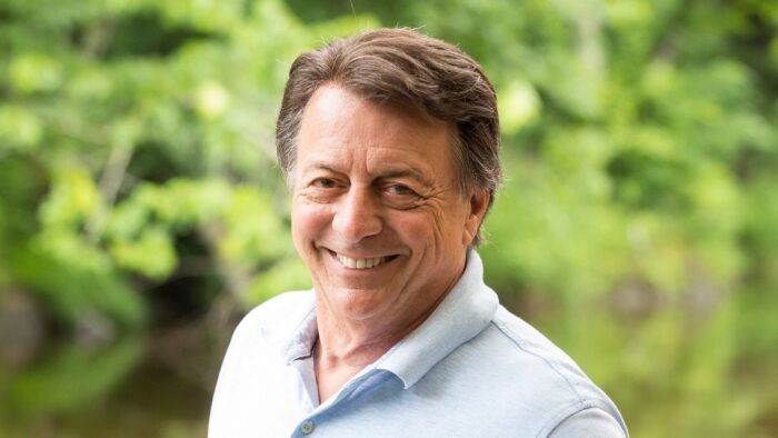 L'animateur Jean-Luc Mongrain, portant un chandail à collet blanc, pose en gros plan, souriant, devant un décor de feuillage vert.