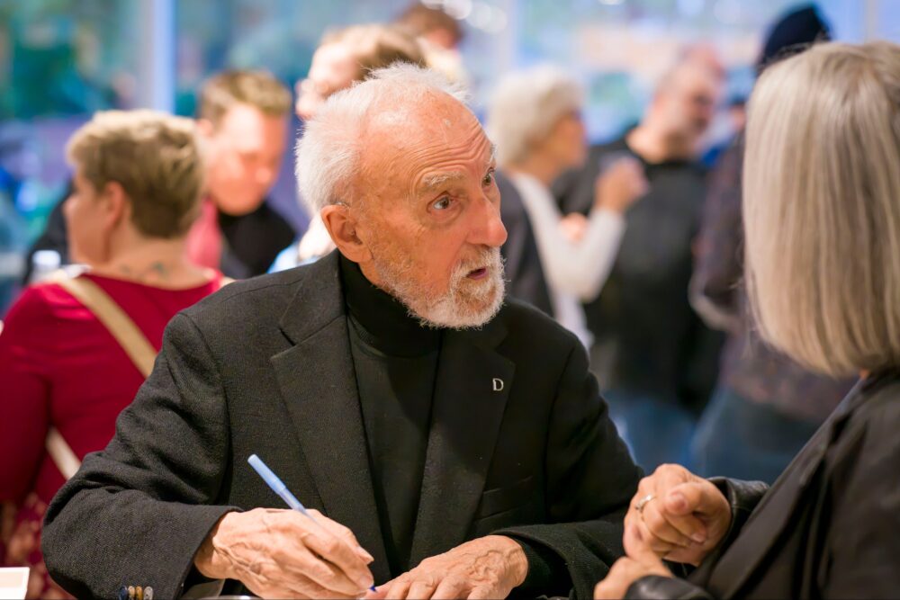 L'humoriste Yvon Deschamps en séance de signature, un crayon à la main, regardant une interlocutrice