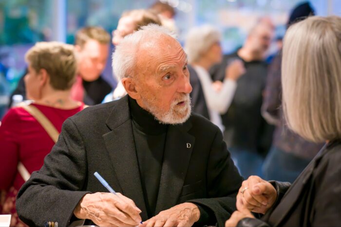 L'humoriste Yvon Deschamps en séance de signature, un crayon à la main, regardant une interlocutrice